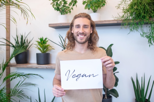 Vegan. Glücklicher junger erwachsener langhaariger Mann, der kleines Plakat mit Wort vegan vor ihm hält