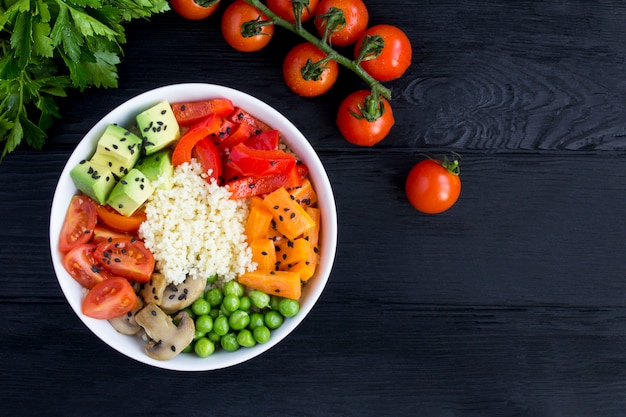 Vegan poke bowl con cuscús y verduras en el tazón blanco sobre el fondo negro de madera. Vista superior. Copia espacio De cerca.