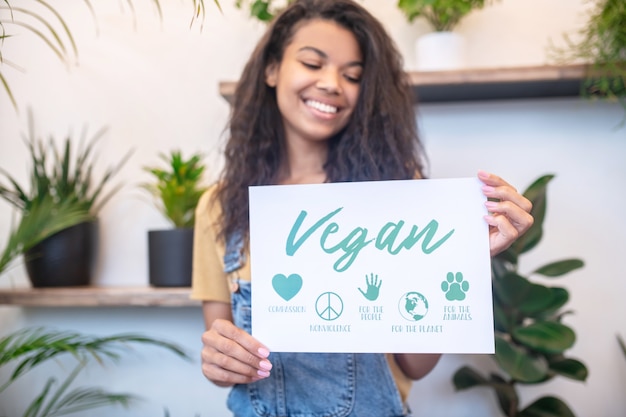 Vegan, sinais. Jovem mulata de cabelos compridos alegre mostrando cartaz com placas veganas em pé dentro de casa