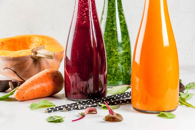 Vegane Diätkost. Bunte frische organische Gemüsesmoothies der Auswahl mit Herbstgemüse: Rote-Bete-Wurzeln, Kürbis, Karotte, Blattgemüse. In Flaschen, mit Strohhalmen, weißer Marmortisch. copyspace