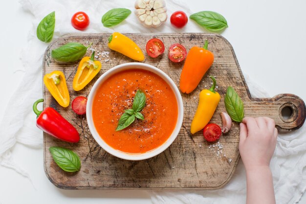 Vegane rote Gemüsesuppe mit Tomaten, Paprika, Knoblauch und Basilikum Kinderhände nehmen Basilikumblatt in der Nähe einer Schüssel mit Tomatensuppe