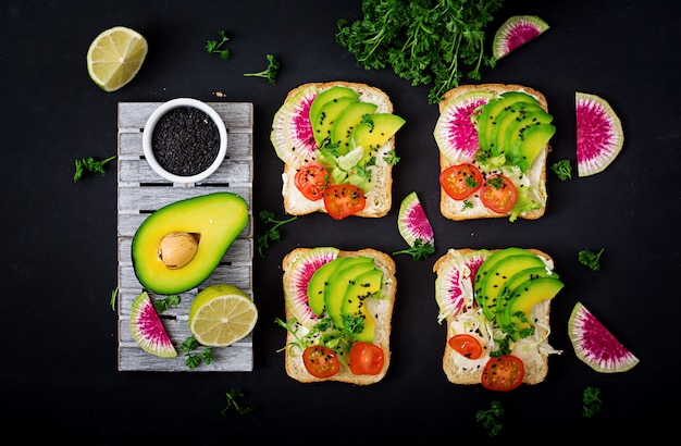 Vegane Sandwiches mit Avocado, Wassermelonenrettich und Tomaten auf einem schwarzen Tisch.