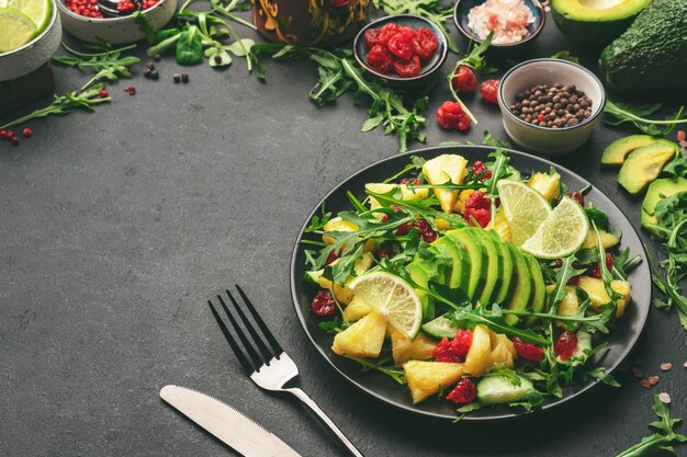 Veganer vegetarischer Salat mit Ananas-Rucola-Avocado und getrockneter Kirsche auf schwarzem Teller auf dunklem Steinküchentisch Draufsicht Konzept zur Gewichtsabnahme bei gesunder Ernährung mit sauberer Ernährung