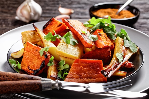 Vegetais de raiz assada marroquinos: nabo e cenoura com especiarias ras el hanout: canela, açafrão, anis servido em um prato com coentro fresco e talheres em um fundo de madeira, vista de cima, close-up