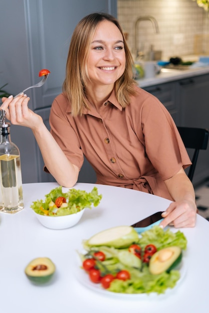 Vegetariana sorridente degustação feminina por colher salada de legumes fresca enquanto está sentado à mesa na cozinha com interior moderno Conceito de alimentação saudável