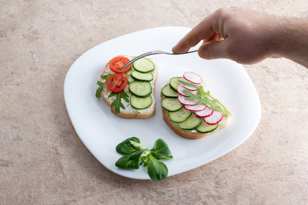 Vegetarische Sandwiches mit Hüttenkäse, Gurken, Tomaten und Radieschen auf einem weißen Teller. Eine Hand hält eine Gabel über einem weißen Teller mit Sandwiches.