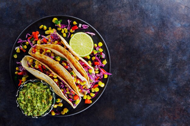 Vegetarische Tacos mit Mais, Purpurkohl und Tomaten auf einem schwarzen Teller. Tacos mit Gemüse und Guacamole-Sauce auf dunklem Hintergrund. Platz kopieren. Ansicht von oben