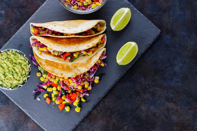 Vegetarische Tacos mit verschiedenem Gemüse, Guacamole und geschnittenem Kalk auf dunklem Hintergrund. Tacos mit Zuckermais, Purpurkohl und Pfeffer auf einem Schieferbrett. Platz kopieren