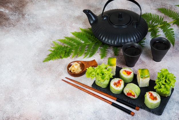 Vegetarische vegetarische Sushi-Rollen mit einer Teekanne Tee und chinesischen Tassen stehen auf einem Betontisch.