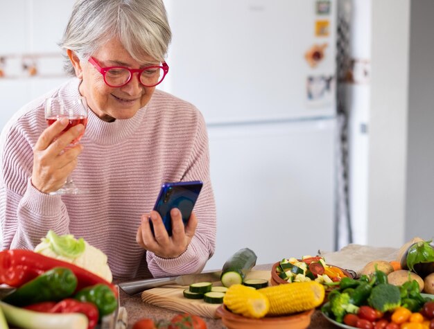 Vegetarischer Lebensstil. Ältere Frau liest eine Nachricht auf dem Smartphone mit einem Rotweinglas in der Hand. Auf dem Tisch Mix aus rohem Saisongemüse