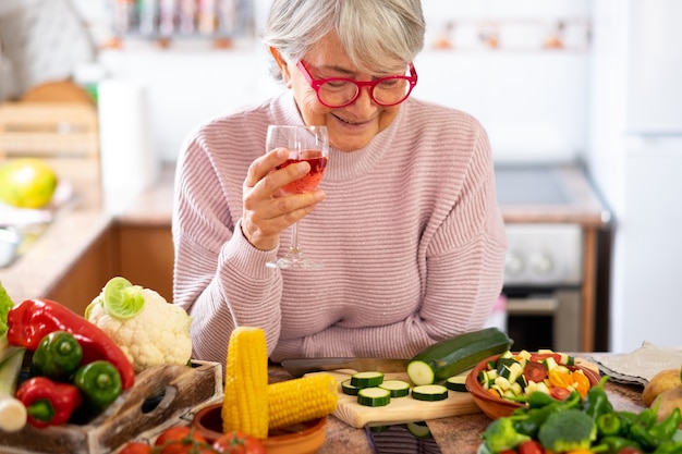 Vegetarischer Lebensstil. Schöne ältere Frau in der Küche mit einem Rotweinglas in der Hand, Blicke auf den Tisch mischen das rohe Gemüse der Saison und denken, wie man sie zubereitet