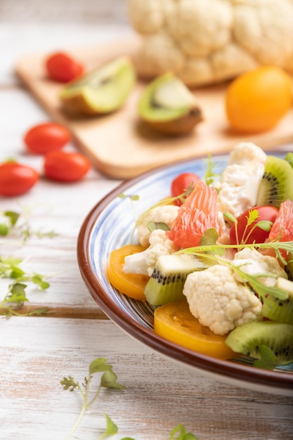 Vegetarischer Salat aus Blumenkohl, Kiwi, Tomaten, Mikrogrünsprossen auf weißem Holzhintergrund und blauem Leinengewebe. Seitenansicht, Nahaufnahme, selektiver Fokus.
