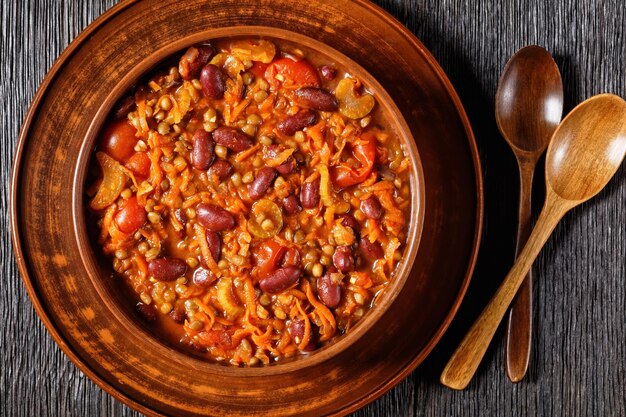 Vegetarisches Chili mit Kidneybohnen und Linsen in Steingutschale auf dunklem Holztisch, horizontale Ansicht von oben, flache Lage