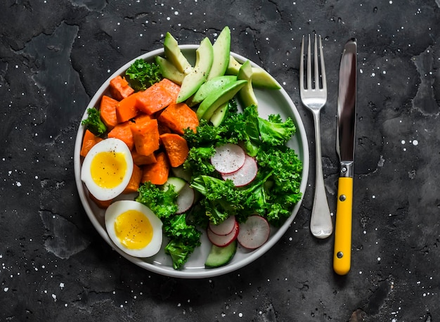 Vegetarisches Mittagessen gekochter Süßkartoffel-Eikohl-Avocado-Rettich-Gurkensalat auf einer Draufsicht des dunklen Hintergrundes