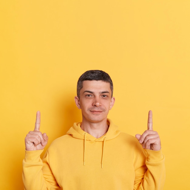 Veja o anúncio Sorrindo, homem calmo e bonito, com cabelo escuro, vestindo um moletom com capuz de estilo casual, isolado sobre fundo amarelo, apontando para o espaço da cópia para cima na área de anúncio