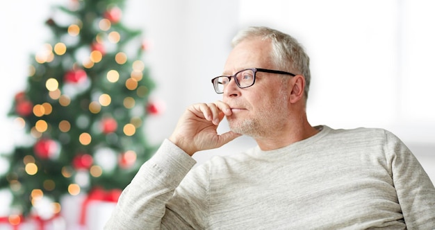 La vejez, las vacaciones, el problema y el concepto de la gente: un anciano con anteojos pensando en casa sobre el fondo del árbol de Navidad