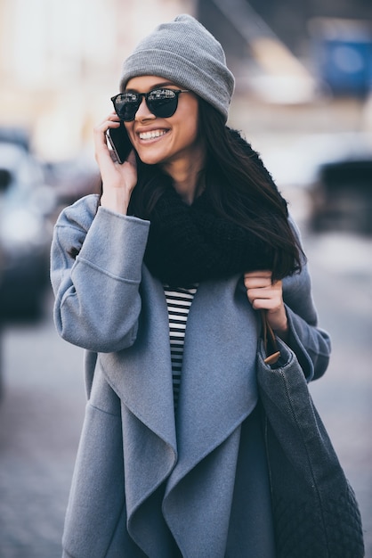 Vejo você em breve! Mulher jovem e bonita em óculos de sol falando no celular e desviando o olhar com um sorriso enquanto caminha ao ar livre