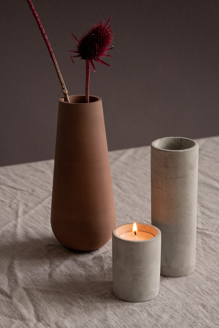 Foto vela acesa, vidro ou vaso de cerâmica branca comprida e jarro de argila marrom com duas flores silvestres secas sobre uma toalha de mesa de linho na parede preta