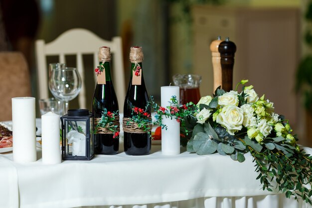 La vela blanca ancha y dos botellas verdes con vino tinto decorado con flores, verdor y cordel