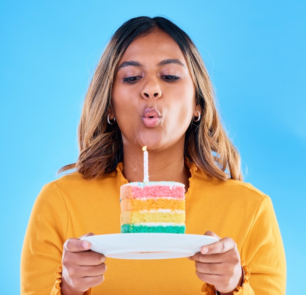 Foto vela de bolo de aniversário soprando e mulher em um estúdio com celebração e sobremesa comida de festa isolada e fundo azul de uma jovem fêmea com guloseima doce e arco-íris com surpresa e prato