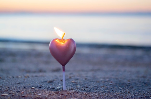 Vela em forma de coração queimando na areia da praia perto das ondas do mar durante o amanhecer do pôr do sol queimando a vela para