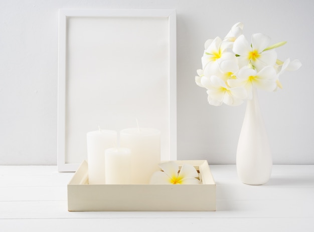 Velas brancas em uma bandeja de madeira branca, moldura de pôster simulada e flores de Plumeria em um vaso de cerâmica moderno sobre o interior da mesa de madeira branca, spa em tom suave natureza morta