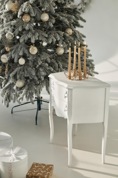 Velas de ouro de Natal acesas queimam em um castiçal na mesa de cabeceira em uma sala decorada com design de Ano Novo perto de abeto de árvore nevado artificial