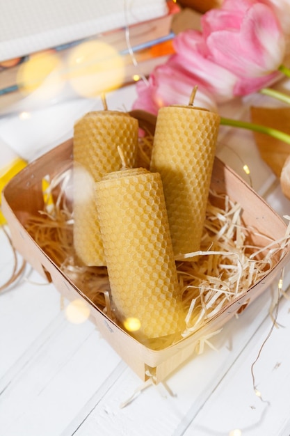 Velas do favo de mel encontram-se em uma cesta perto de tulipas cor de rosa, livros e fairylights no fundo de uma mesa de madeira.