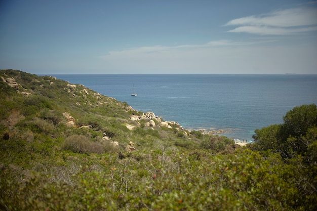 Veleiro sai do mar Mediterrâneo filtrado por um promontório verde com vegetação típica