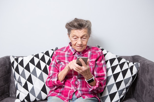 velha caucasiana grisalho com rugas senta-se em casa na sala de estar no sofá e usa telefone celular