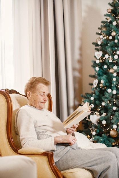 Velha sentada em uma cadeira no dia de Natal. Avó lendo um livro. Mulher vestindo roupas bege.