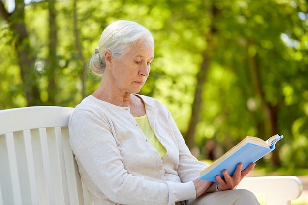 velhice, aposentadoria e conceito de pessoas - mulher idosa feliz lendo um livro sentada em um banco no parque de verão