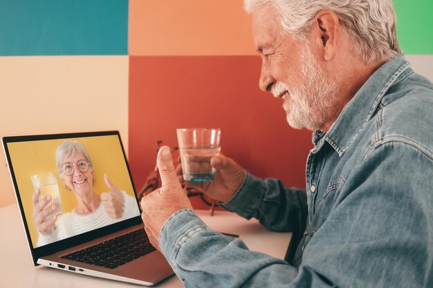 Velho barbudo sorridente conversando em vídeo on-line usando laptop segurando um copo de água