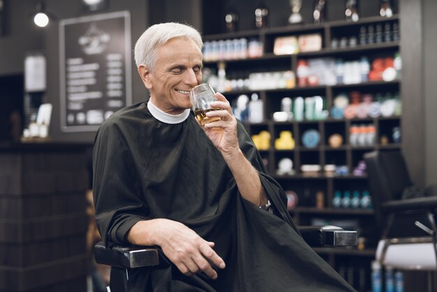 Velho bebe álcool na cadeira do barbeiro na barbearia.