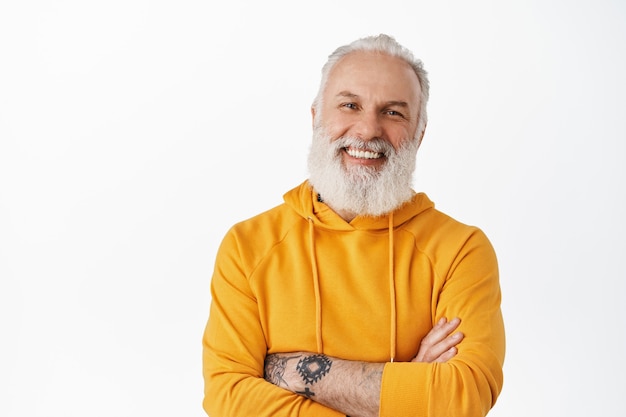 Velho bonito com tatuagens rindo e sorrindo cândido, mostrando emoções autênticas, braços cruzados no peito, de pé relaxado contra uma parede branca