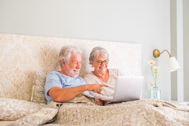 Velho casal caucasiano sênior sorrindo e usando o laptop pela manhã na cama no quarto em casa. casal de idosos navegando e usando mídias sociais no laptop em casa.