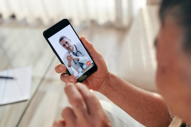 Velho feliz conhecer seu médico por videochamada por telefone inteligente em casa, durante o intervalo de covid-19.