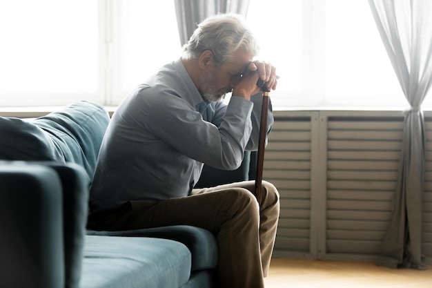 Velho infeliz deprimido sente-se no sofá em casa, segure a vara de madeira, sinta-se abandonado, triste, maduro, homem dos anos 60, com bengala, sofre de solidão ou depressão, conceito de solidão idosa