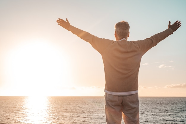 Foto velho olhando para o pôr do sol e o mar de braços abertos, sentindo-se livre e feliz - liberdade e felicidade, estilo de vida e conceito