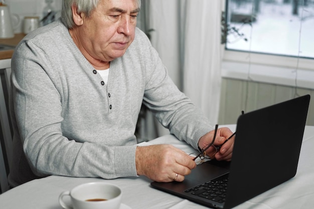 Velho pensando em laptop pensativo maduro cabelos grisalhos anos 60 anos 70 homem sentado à mesa com laptop pensativo aposentado homem sênior trabalha online no computador de casa resolvendo problemas com soluções