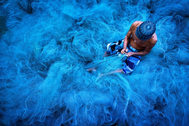 Velho pescador costurando redes de pesca azuis sentado no chão