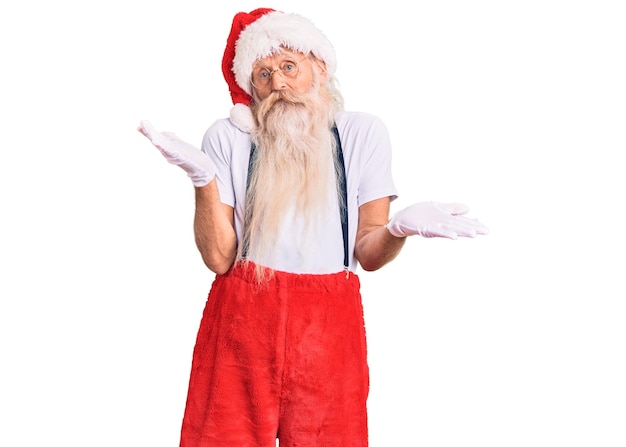 Velho sênior com cabelos grisalhos e barba longa vestindo fantasia de Papai Noel com suspensórios sem noção e expressão confusa com os braços e as mãos levantadas. conceito de dúvida.