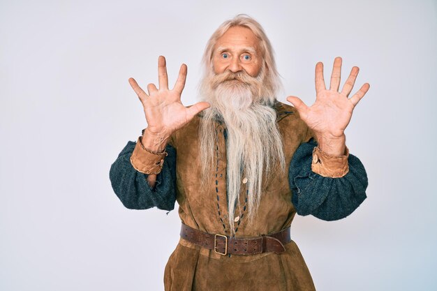 Velho sênior com cabelos grisalhos e barba longa vestindo traje tradicional viking mostrando e apontando para cima com os dedos número dez enquanto sorria confiante e feliz
