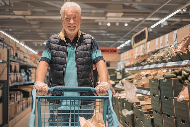 Velho sênior empurrando o carrinho de compras no supermercado comprando mantimentos tendo cuidado com o preço