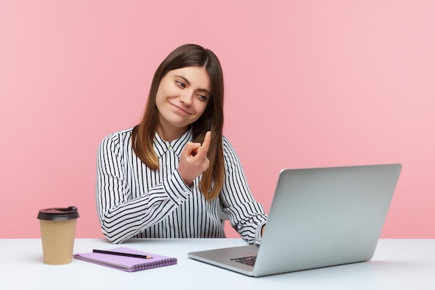 Ven aquí. Hermosa mujer positiva haciendo un gesto de señas con el dedo hablando en una videollamada usando una laptop, teniendo una comunicación romántica en línea. Disparo de estudio interior aislado sobre fondo rosa