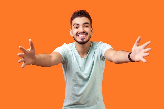 Ven a mis brazos Retrato de un hombre moreno extremadamente feliz y afable con barba en una camiseta blanca que se acerca a la cámara estirando los brazos para abrazarte, una foto de estudio aislada en un fondo naranja