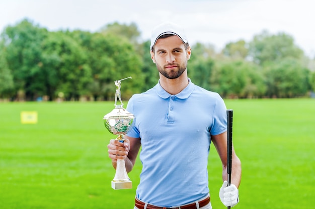 Vencedor confiante. Jogador de golfe confiante segurando a bola de golfe e o driver enquanto está de pé no campo de golfe e olhando por cima do ombro