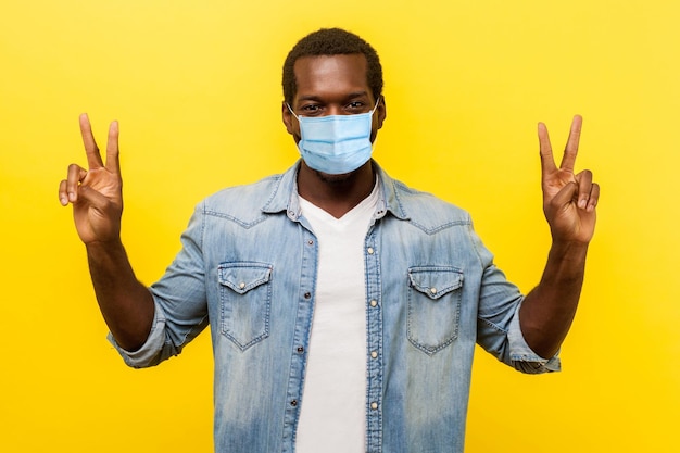 Vencedor de sucesso Retrato de homem animado com sorriso com máscara médica cirúrgica em pé com as mãos levantadas e mostrando sinal de v ou tiro de estúdio de gesto de vitória de paz isolado em fundo amarelo