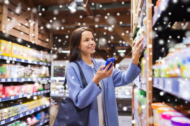 Foto venda, compras, consumismo e conceito de pessoas - jovem feliz com smartphone escolhendo e comprando comida no mercado sobre efeito de neve