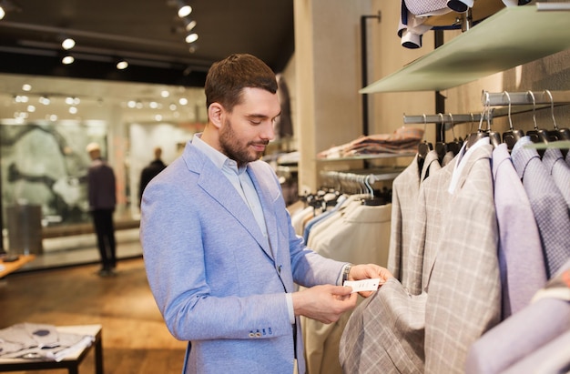 venda, compras, moda, estilo e conceito de pessoas - jovem elegante em jaqueta escolhendo roupas e olhando a etiqueta de preço no shopping ou loja de roupas